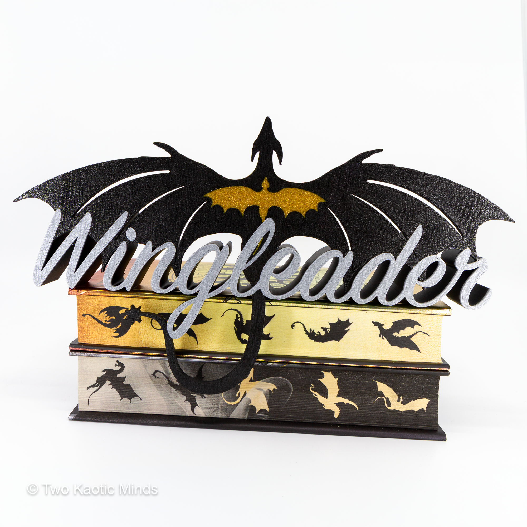 3D printed Wingleader shelf sign in black, gold, and silver with Wingleader script and a spread-wing dragon perched on top, tail curling down over a stack of books, front angle view