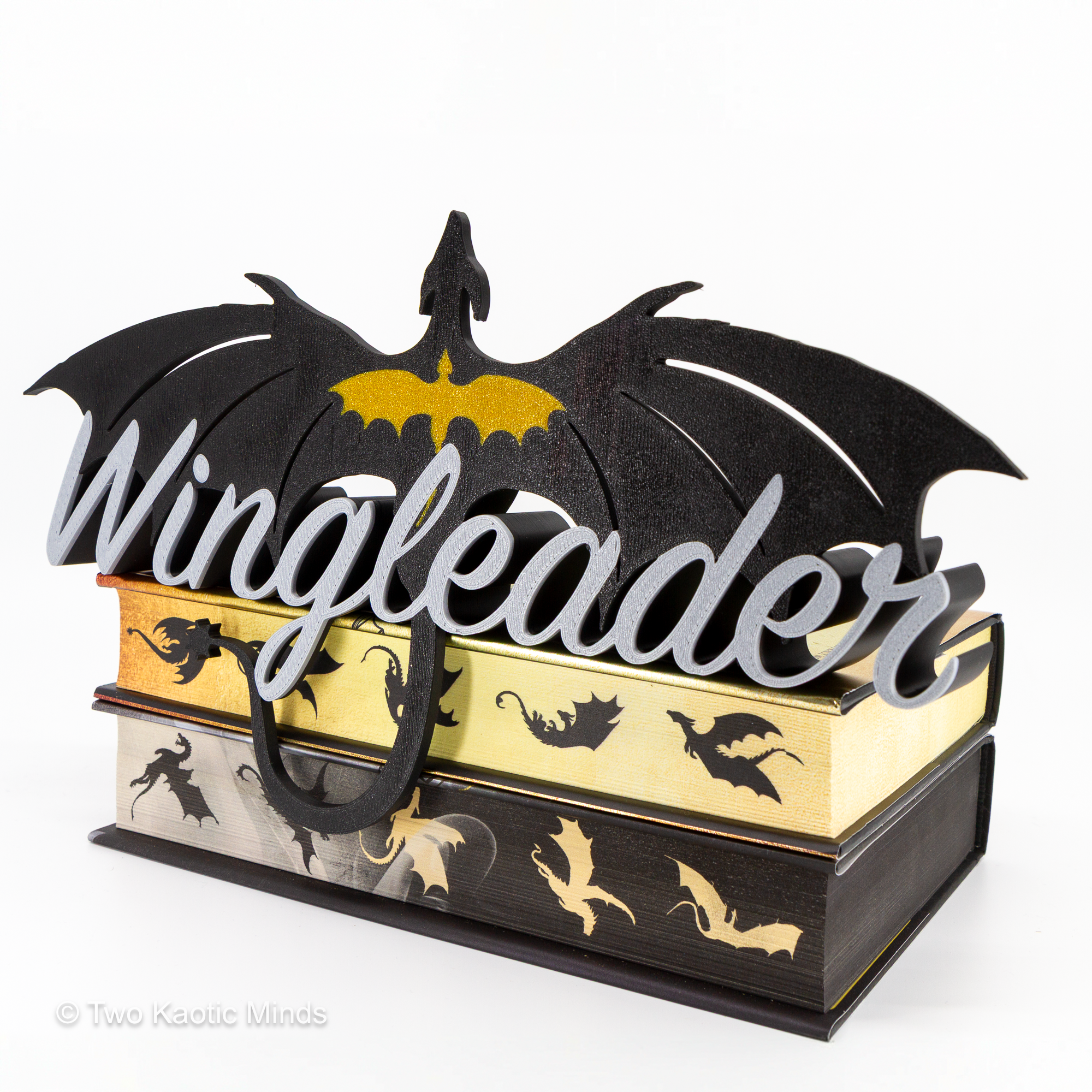 Angled right-side view of the 3D printed Wingleader shelf sign in black, gold, and silver showing the dragon topper and the sign's depth as it sits on a stack of books
