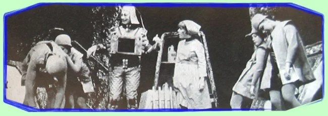 Black and white photograph of children dressed in scout uniforms, standing outdoors beside a wooden fence.