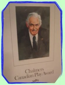 Photograph of an elderly man with white hair, dressed in a dark suit and tie, displayed in a frame. The frame has a sign reading 'Chalmers Canadian Play Award 1985'.
