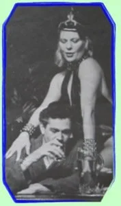 Black and white photo of a woman standing behind a man sitting at a table, both dressed in vintage clothing, with drinks on the table.