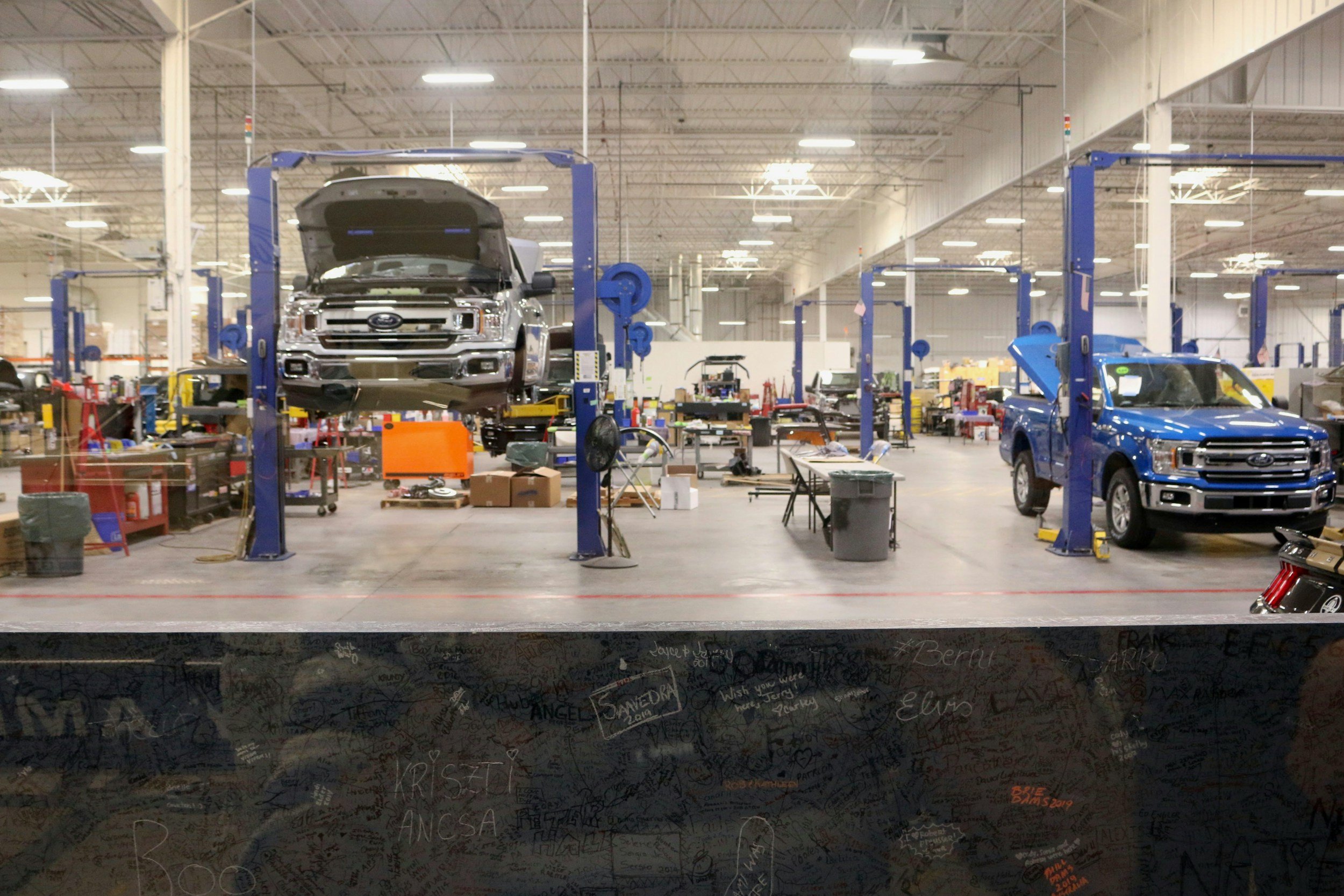 An automotive repair shop with two Ford vehicles on hydraulic lifts, one with an open hood, within a brightly lit warehouse filled with tools and equipment.