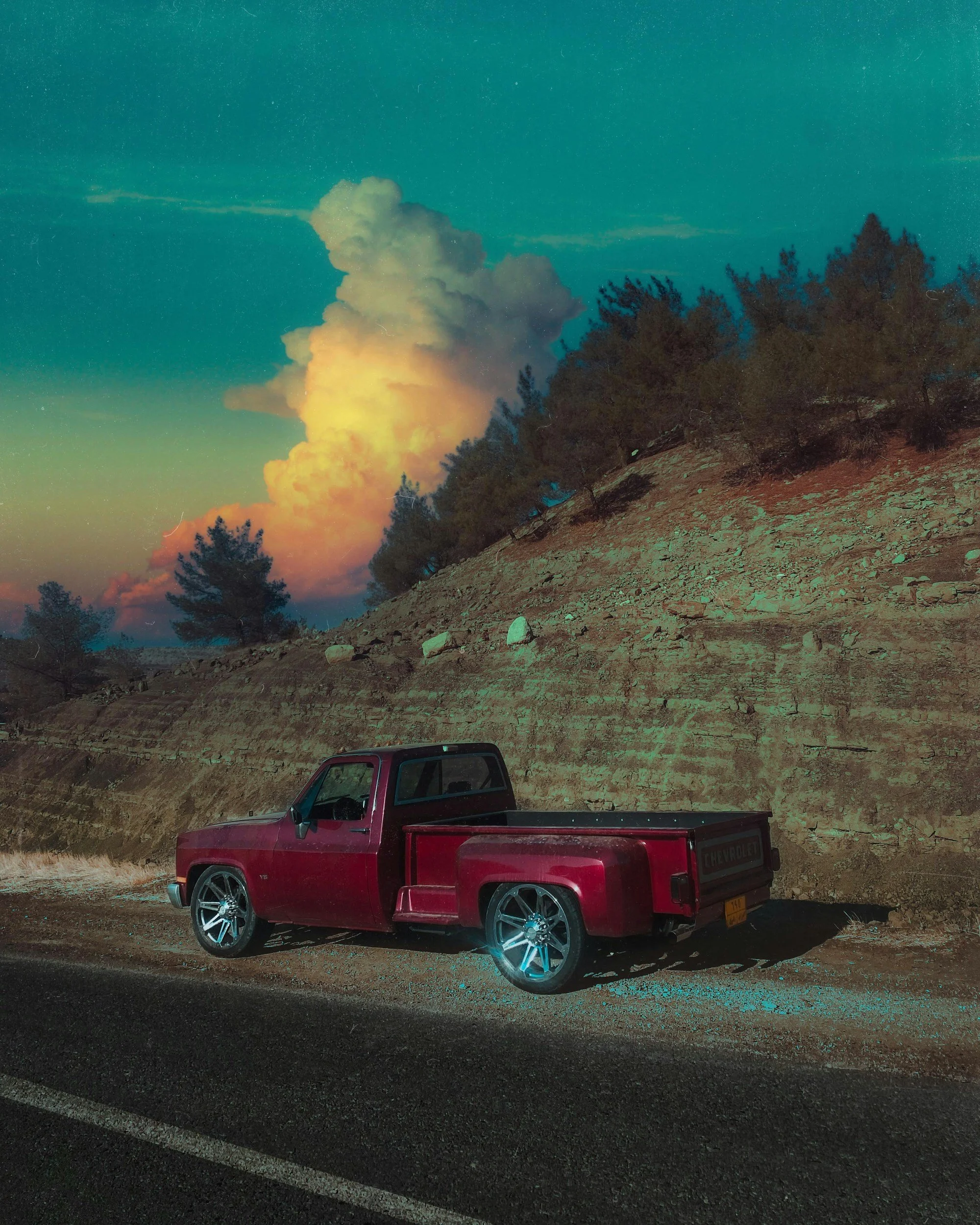 A red Chevrolet Silverado pickup truck parked on the side of a road during sunset, with a smokey eruption or large cloud of smoke in the background, and trees on a hillside.