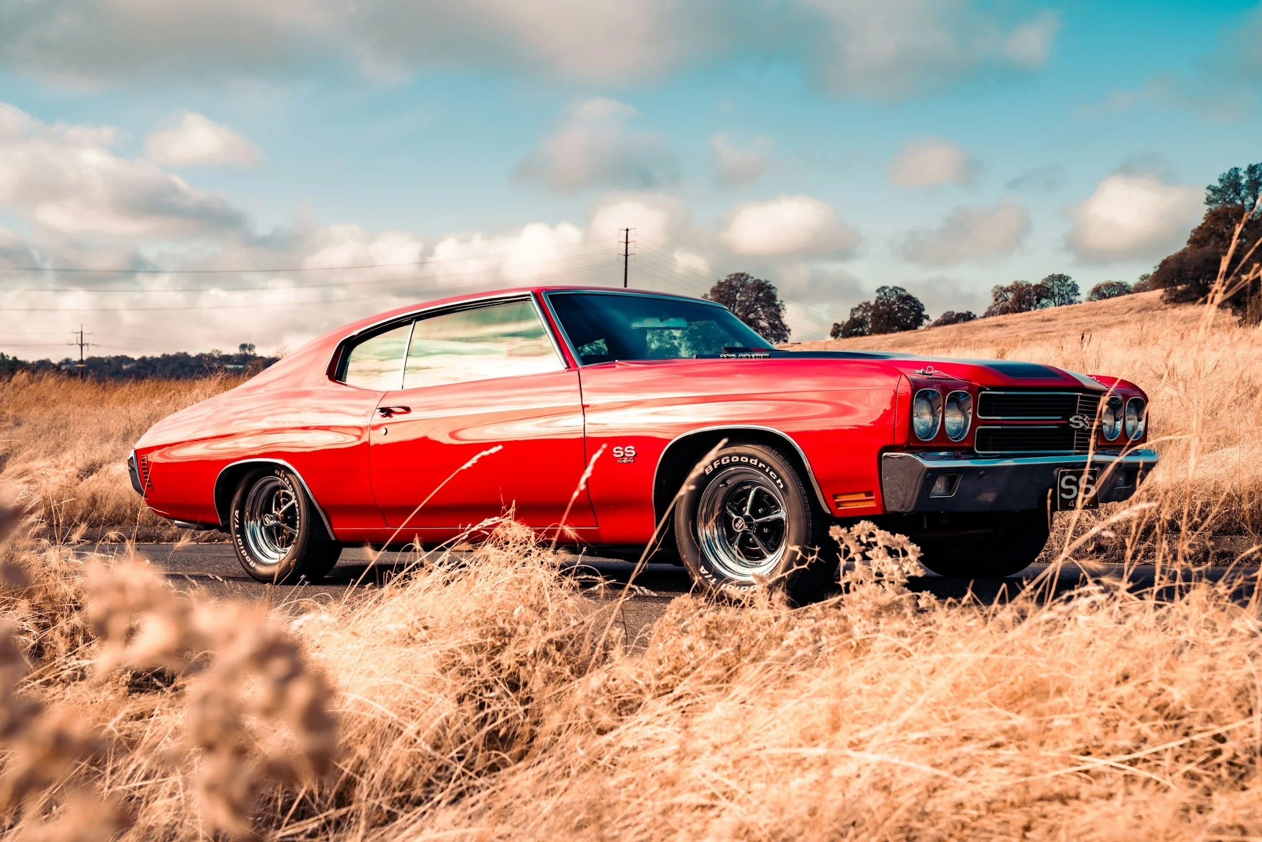 A vintage red Chevrolet SS muscle car parked on a rural road surrounded by tall, dry grass under a partly cloudy sky.