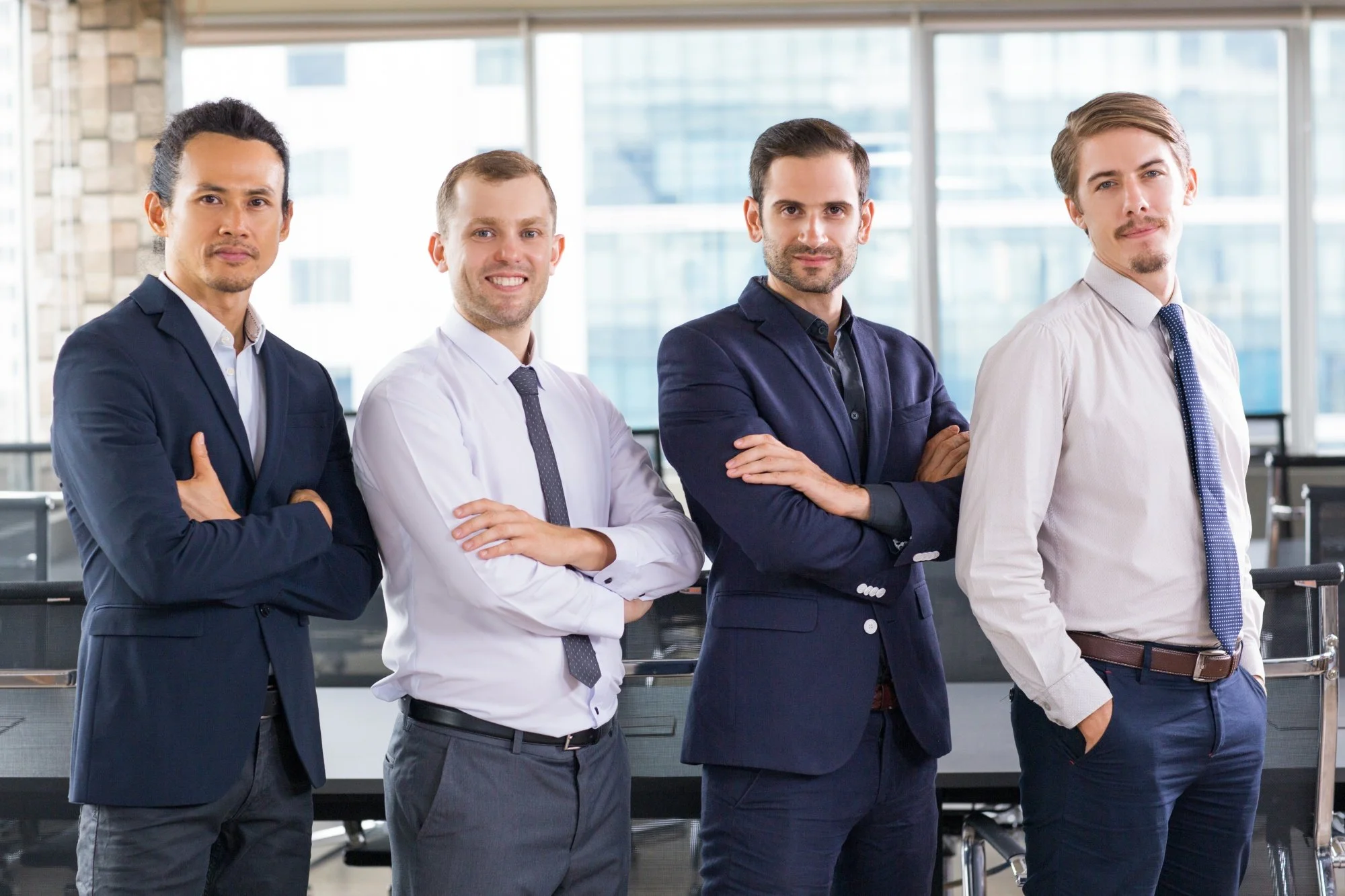 Four professionally dressed men standing with arms crossed in an office with large windows and modern buildings outside.