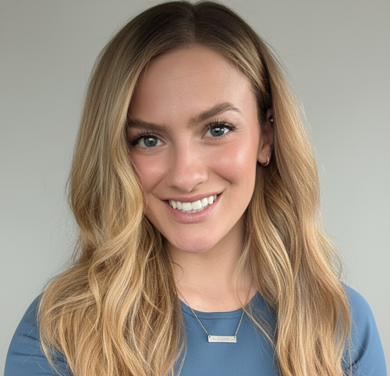 Young woman with long wavy blonde hair, blue eyes, wearing a blue top, silver jewelry, smiling, neutral background.