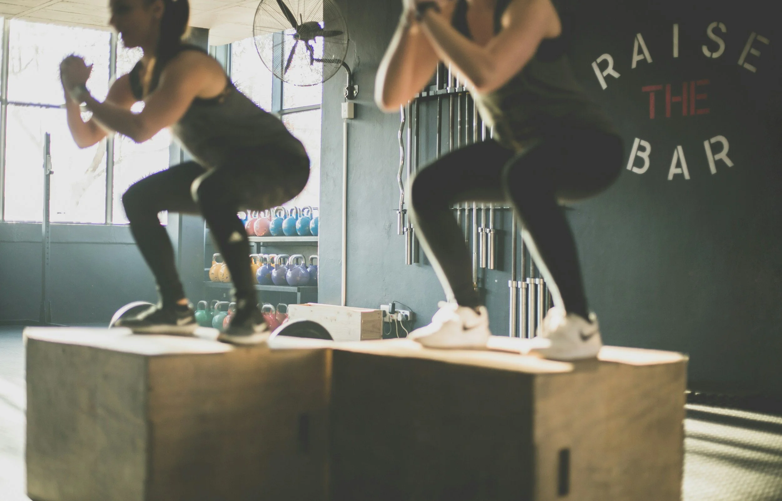 Two women are doing box jumps on a wooden platform at a gym. They are wearing workout clothes and sneakers, with one in a gray tank top and the other in a black top. The gym has kettlebells on shelves in the background and a large fan on the wall.