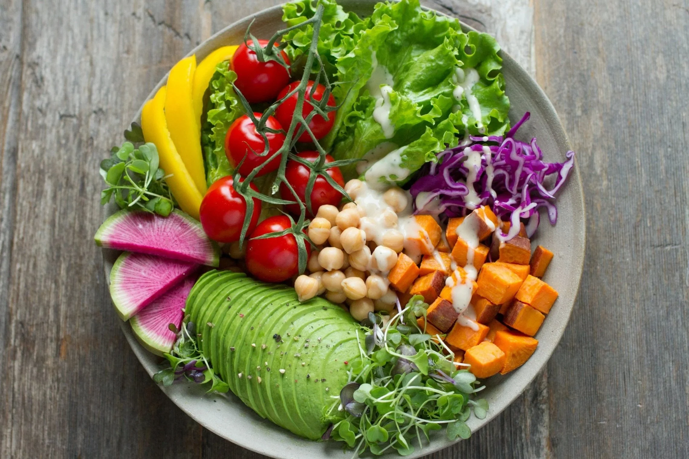 A bowl of fresh salad with cherry tomatoes, sliced avocado, shredded purple cabbage, chopped sweet potatoes, chickpeas, sliced radish, mixed greens, and dressing, on a rustic wooden surface.