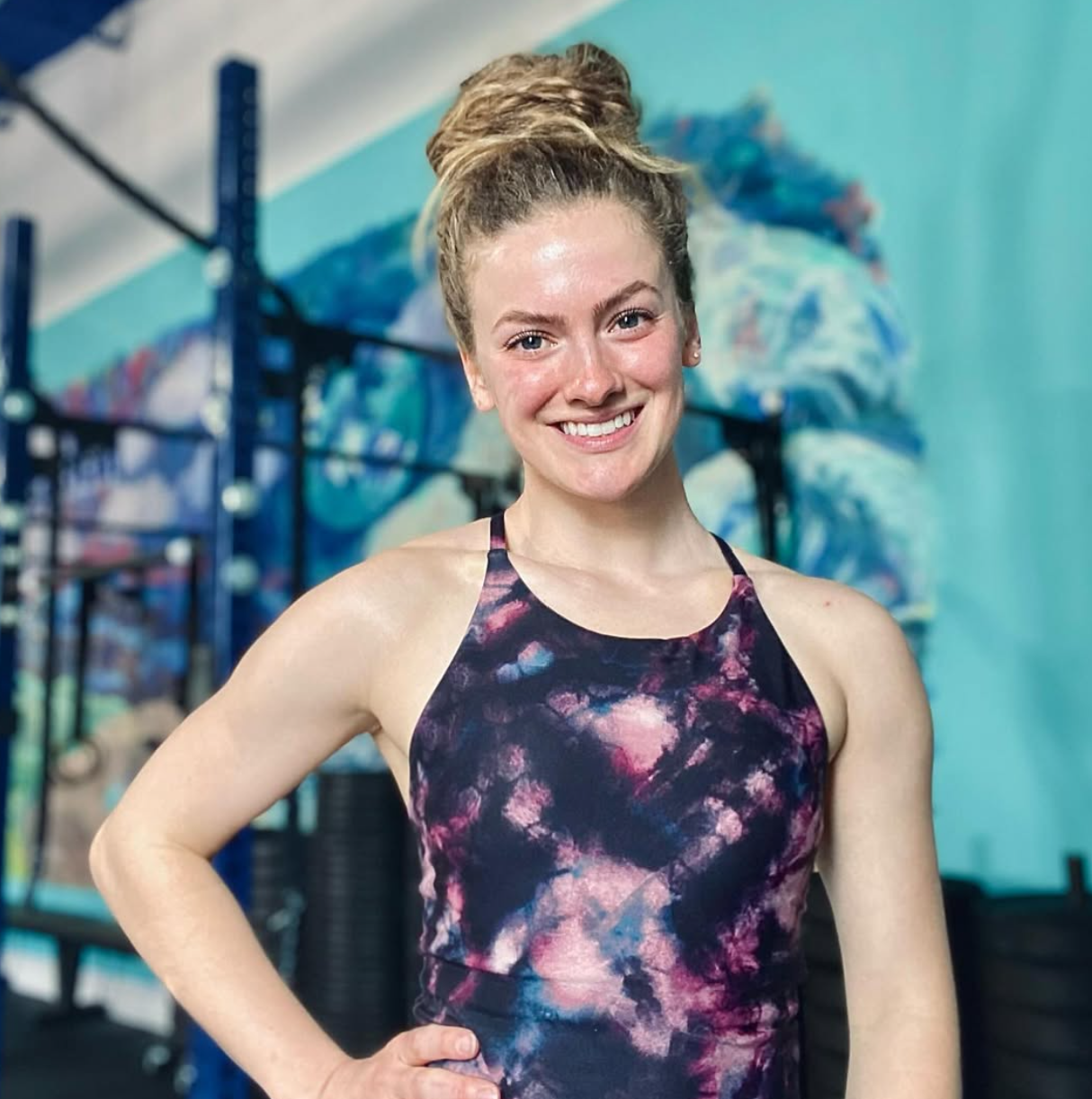 A smiling young woman with a high bun hairstyle, wearing a colorful tie-dye athletic top, standing in a gym with workout equipment and a blue wall in the background.
