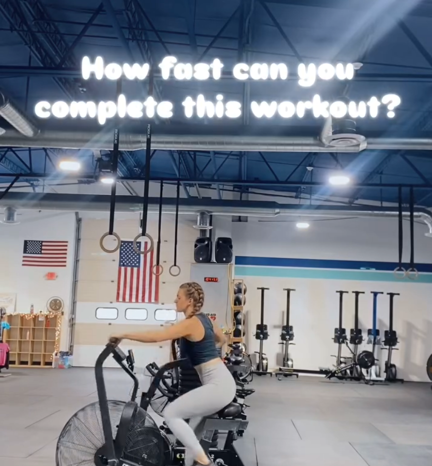 Woman on a stationary exercise bike inside a gym with gym equipment and American flags in the background, illuminated by a digital sign that asks 'How fast can you complete this workout?'