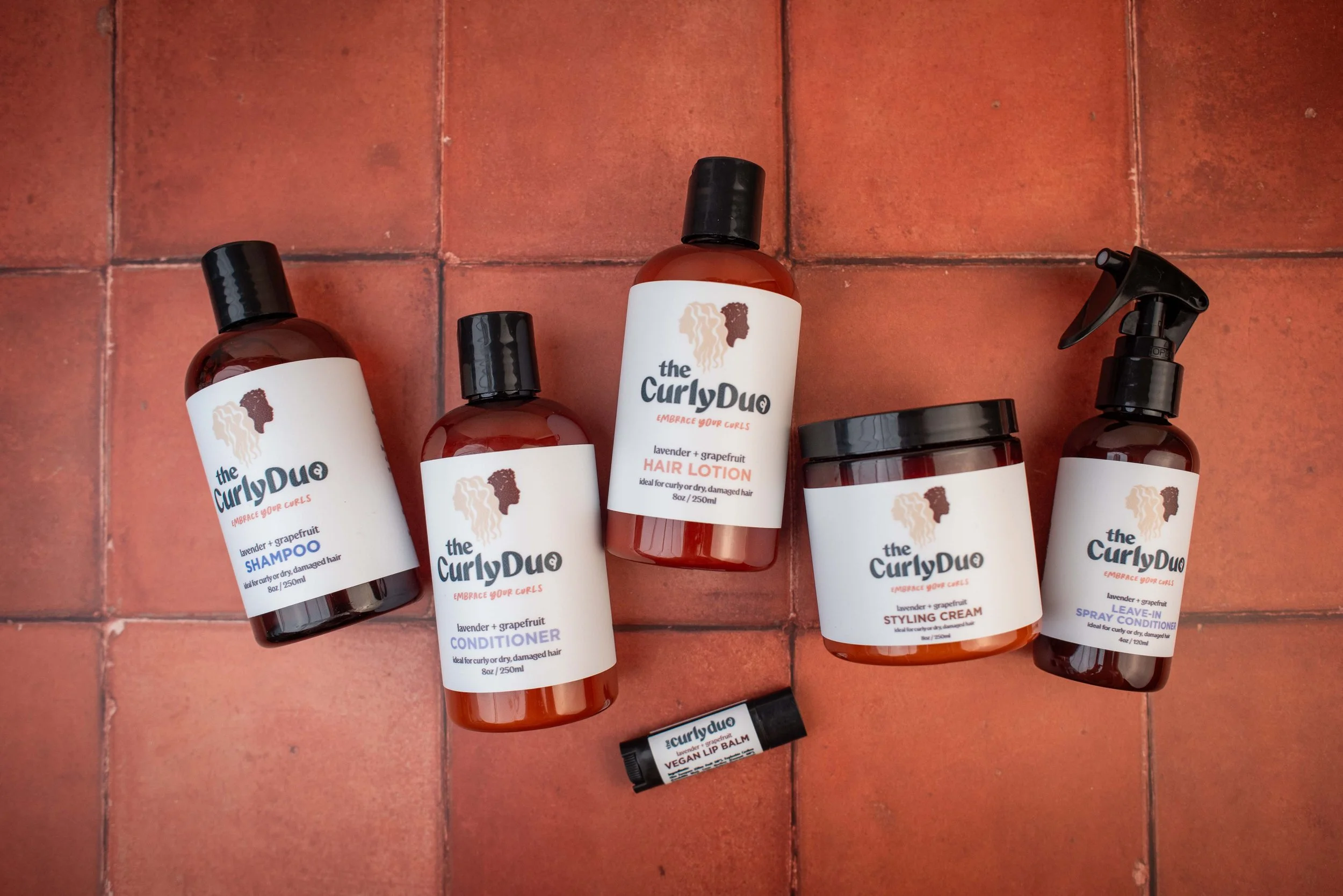 Six hair care products from the Curly Duo brand are arranged on a reddish-brown tiled floor. The products include shampoo, conditioner, hair lotion, styling cream, leave-in spray, and a vegan lip balm.