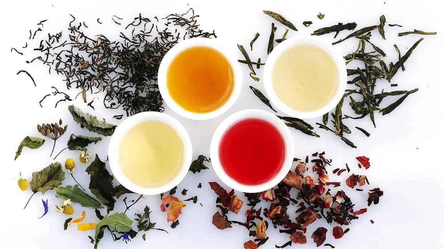 Four cups of different colored teas arranged in a diamond shape on a white surface, surrounded by various dried herbs, flowers, and tea leaves.
