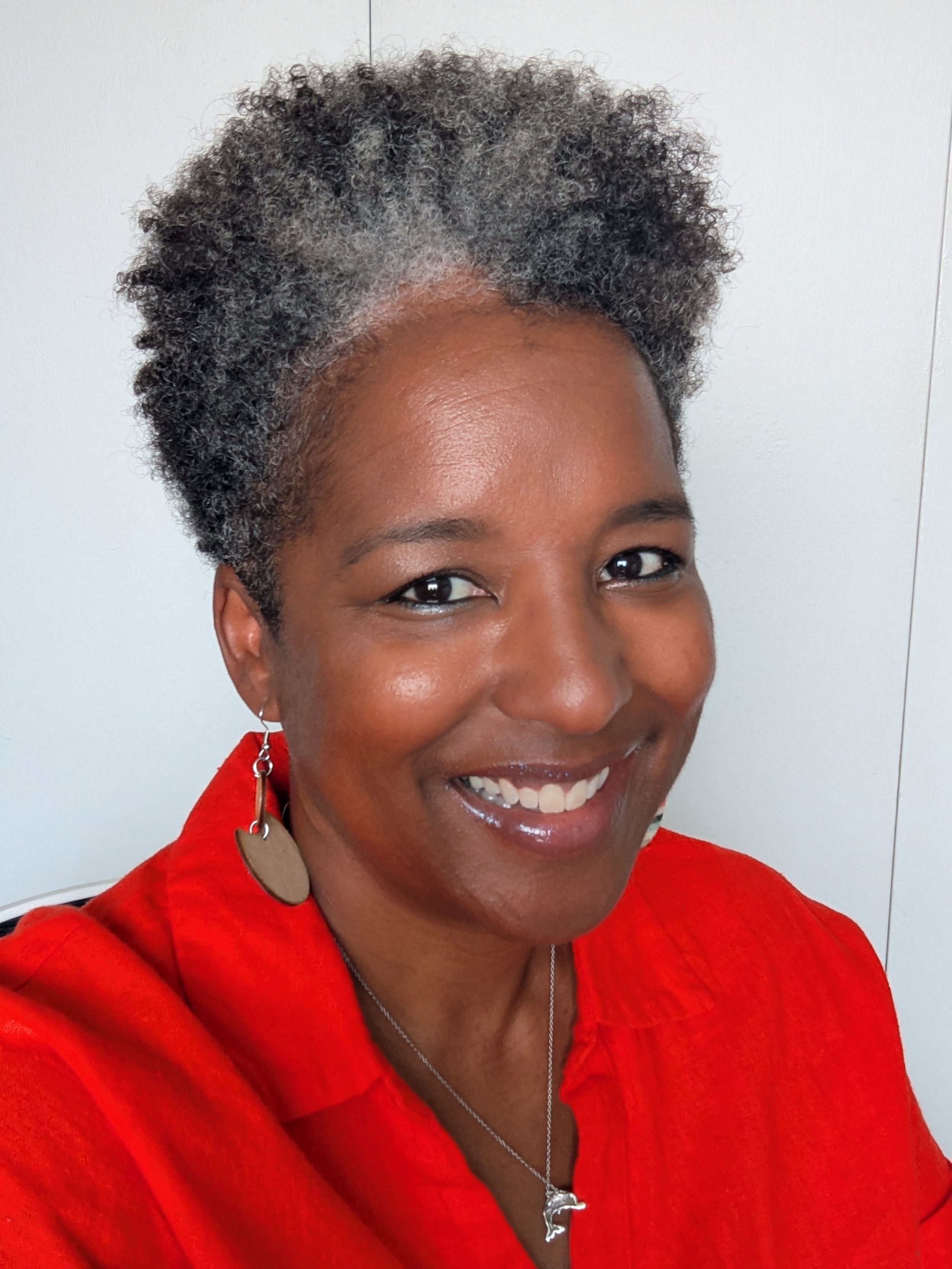 A smiling woman with short, curly gray and black hair, wearing a red blouse, earrings, and a necklace, against a white background.