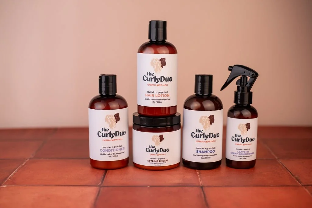 Collection of hair care products from the brand 'the Curly Duo' including conditioner, styling cream, shampoo, leave-in spray conditioner, and hair lotion, all placed on a wooden surface against a neutral background.