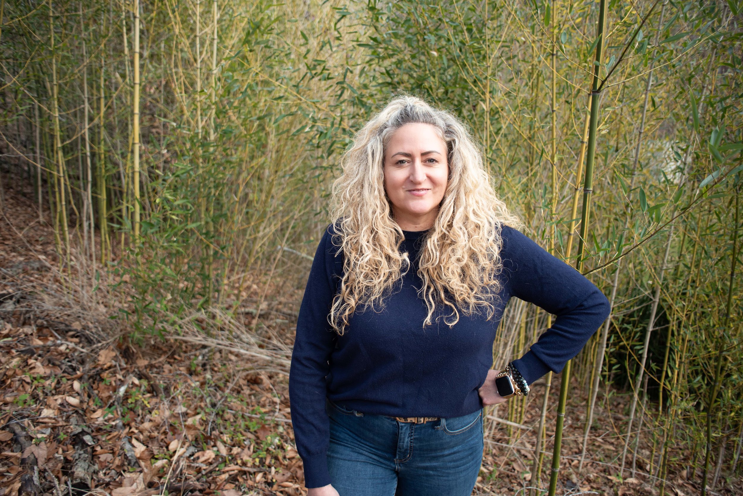 A woman with long curly blonde hair standing in a wooded area with tall green bamboo plants, wearing a navy sweater and blue jeans.