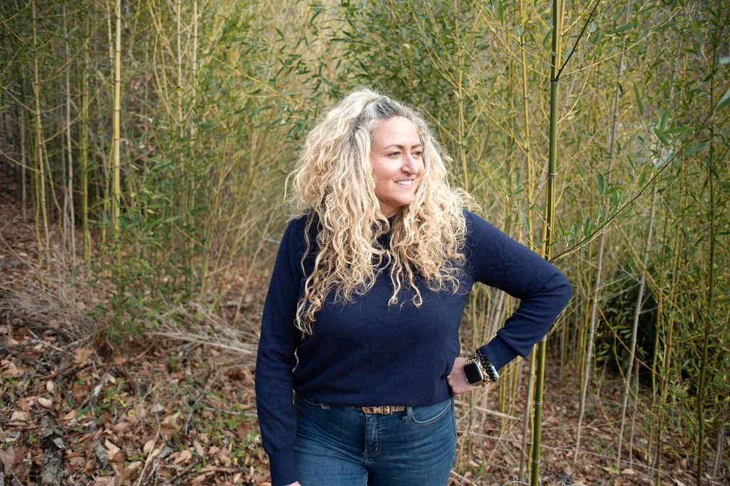 A woman with long, curly blonde hair standing in a bamboo forest, smiling and looking to her right.