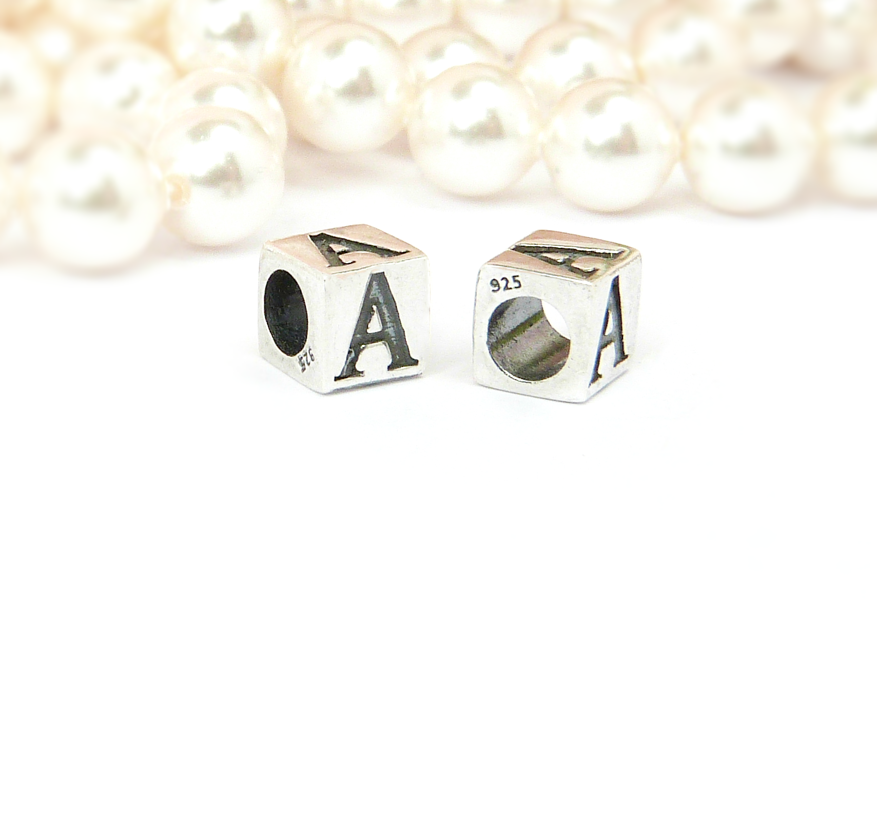 Two sterling silver 925 stamped letter dice beads displaying 'A' with holes on the sides, positioned in front of a string of white pearls.