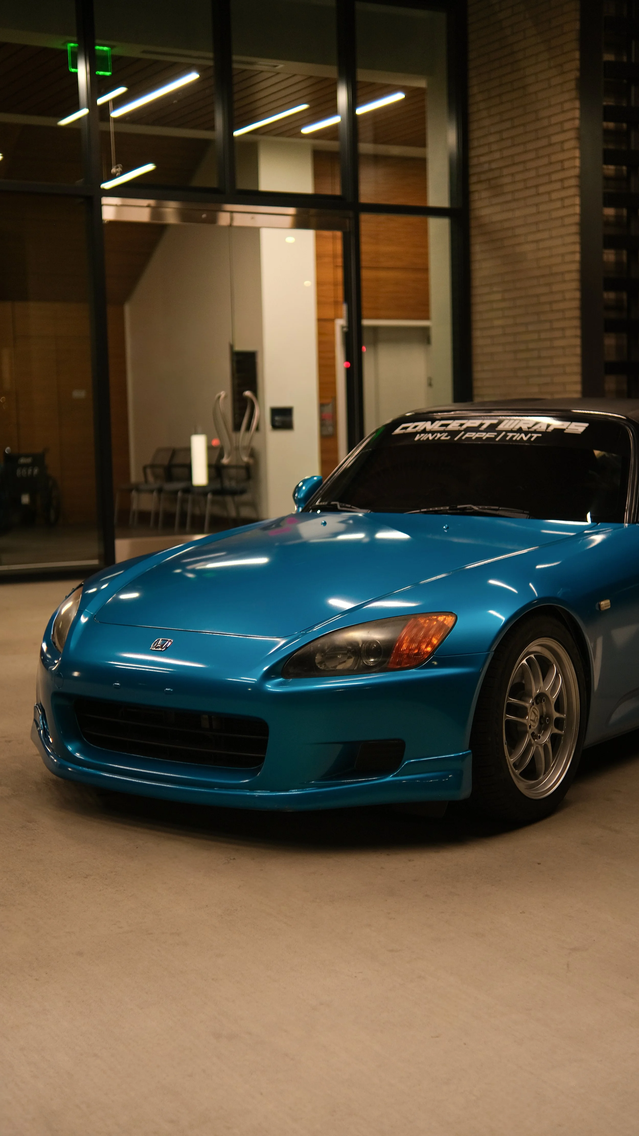 Blue Honda S2000 sports car parked indoors at night, with a building and sitting area visible behind glass doors.