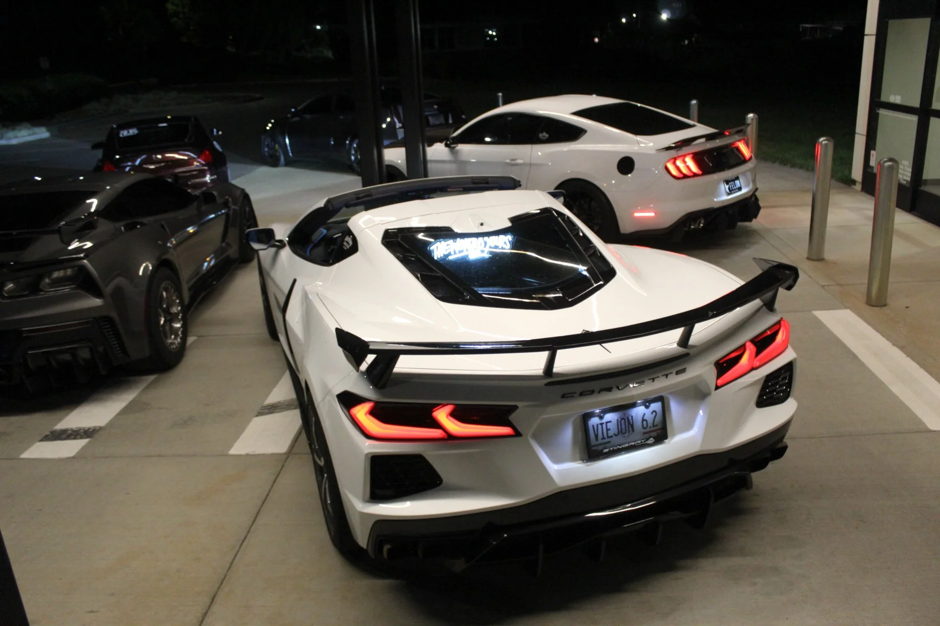 A white Corvette sports car with black accents parked at night, surrounded by other cars including a black sports car to the left and a silver Mustang in the background. The Corvette has bright red tail lights, a black rear spoiler, and a custom license plate that reads 'VIEJON 6.2'.