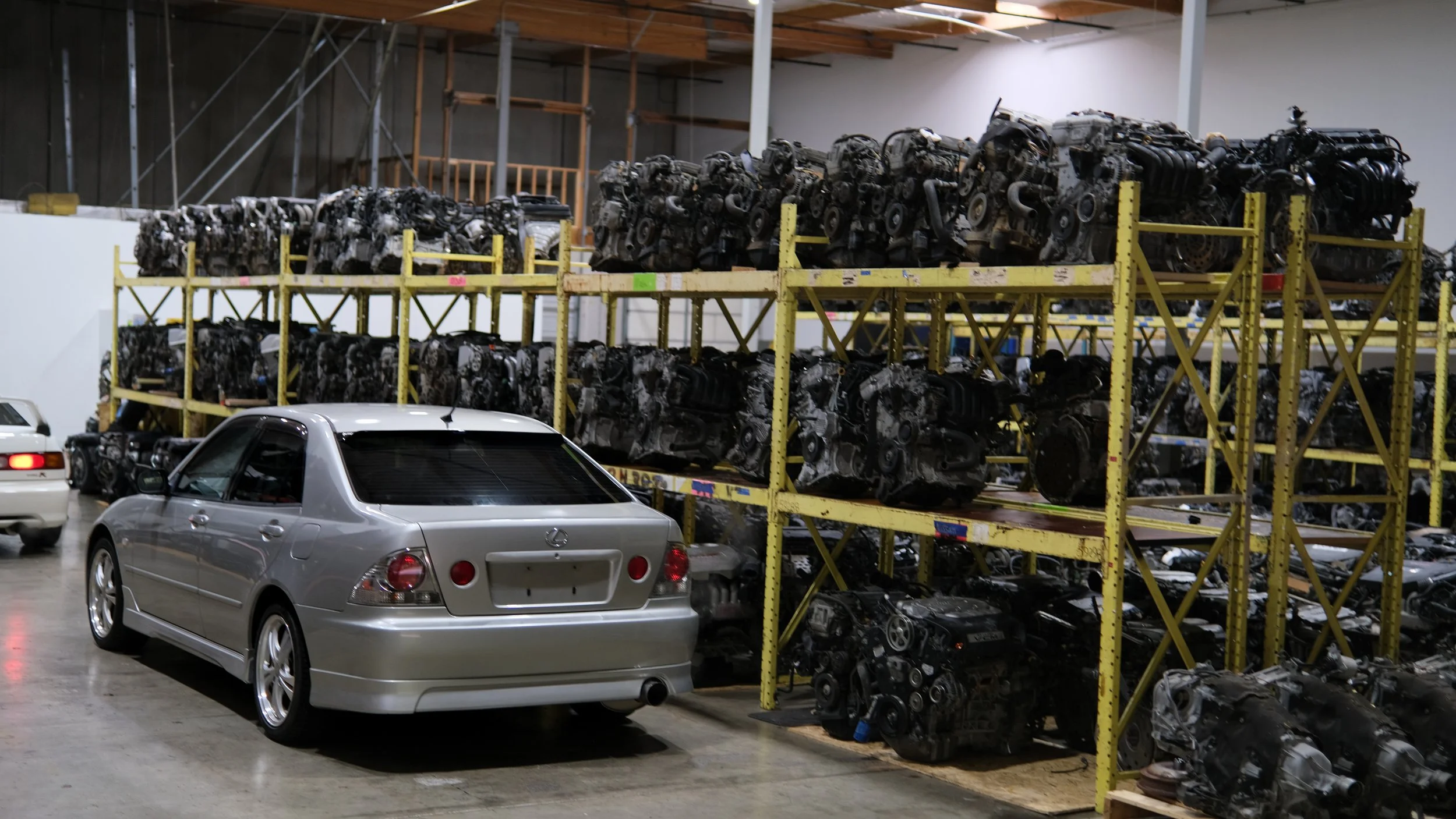 A silver sedan parked in a warehouse with rows of car engines stored on yellow metal racks.