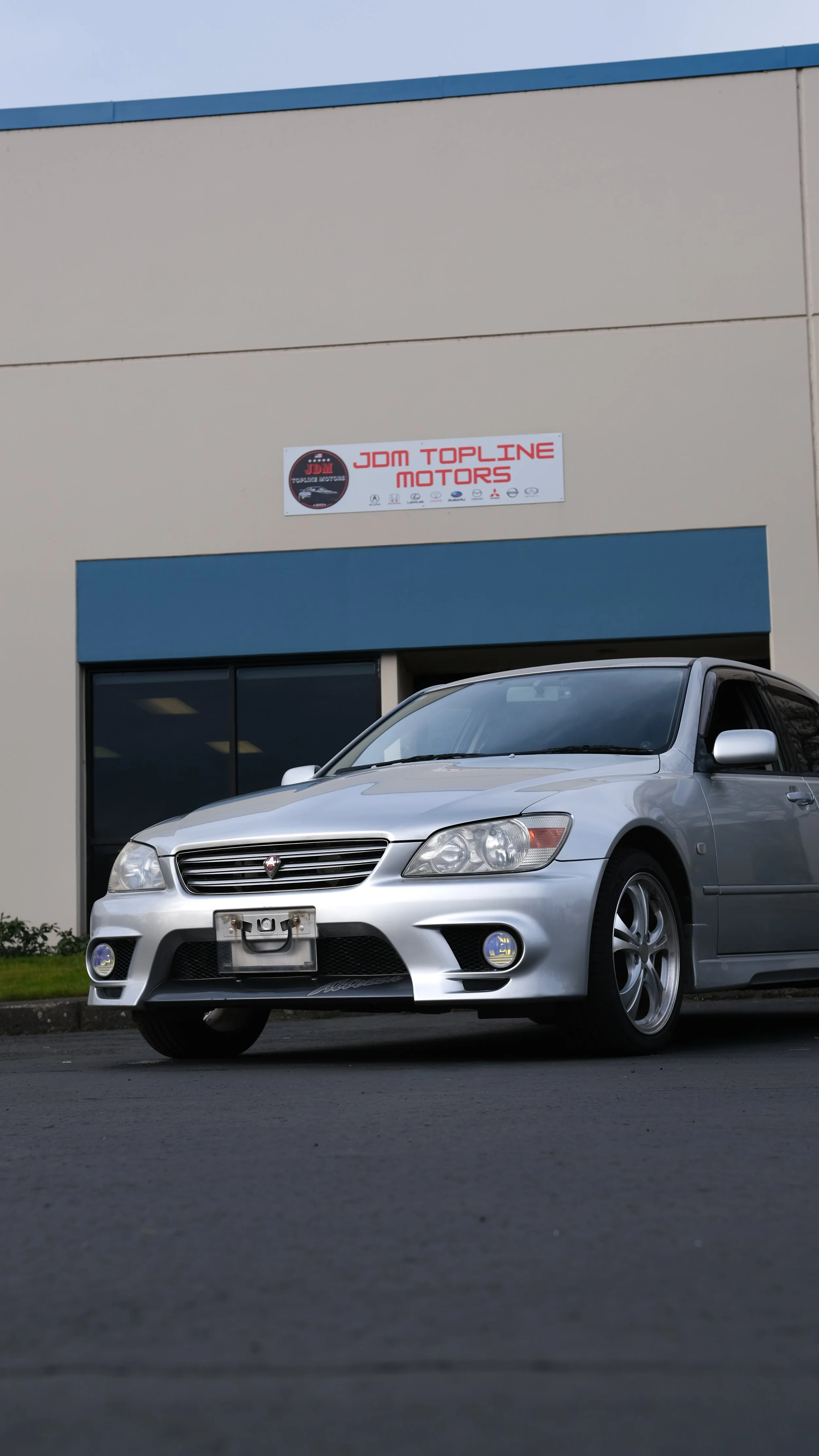 A silver sedan parked in front of a building with a sign that reads 'JDM Topline Motors'.