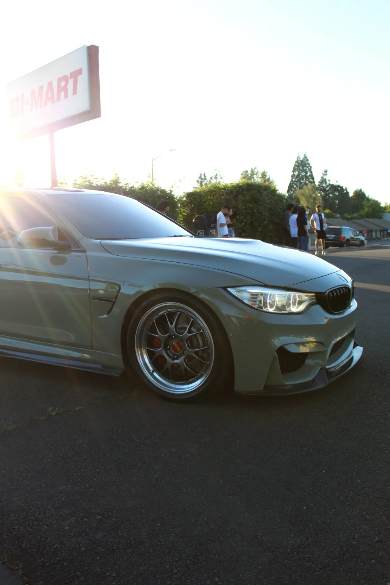 Gray sports car parked in a parking lot near a Bi-Mart store, with people standing in the background and the sun setting.