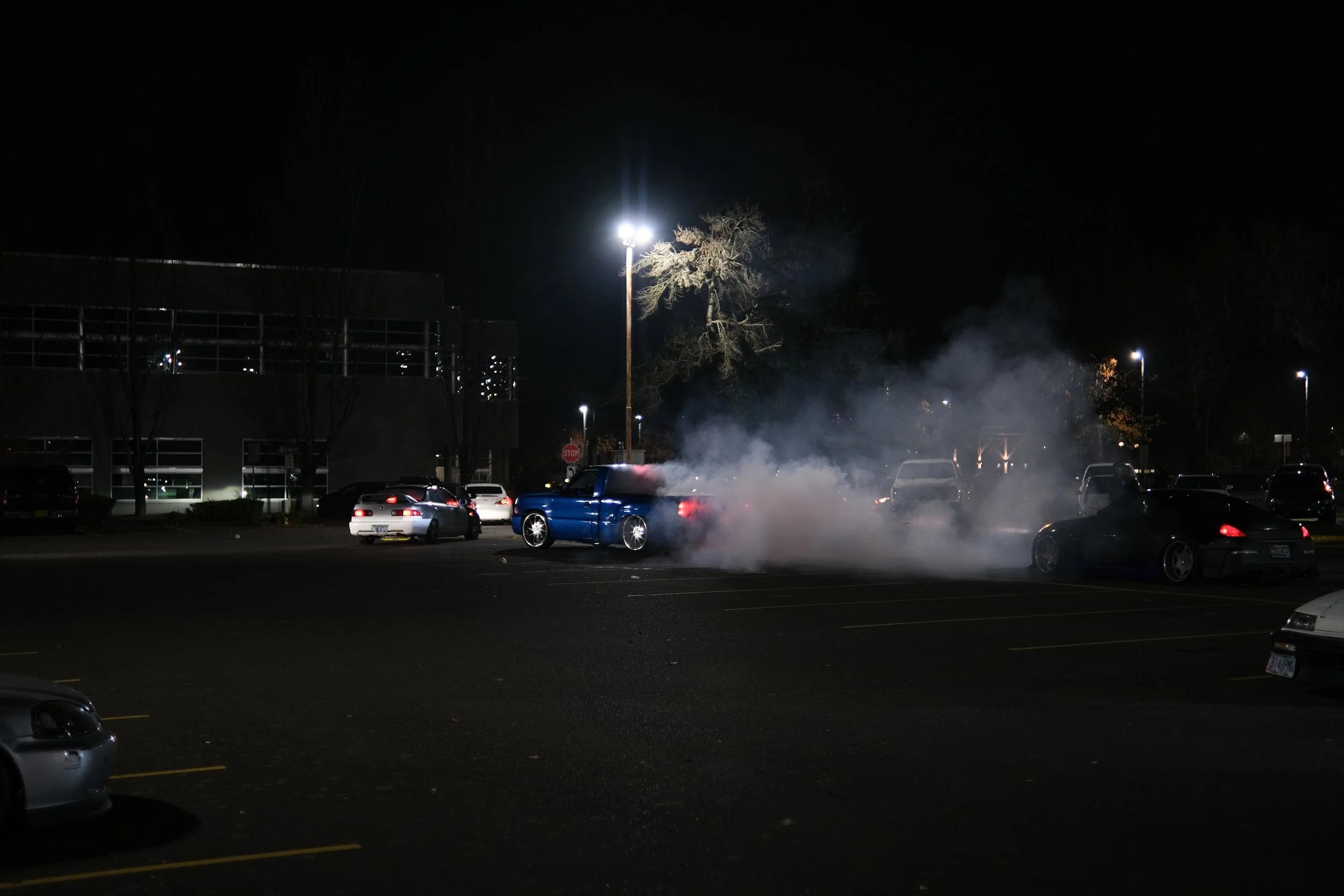 Car emitting white smoke in a dimly lit parking lot at night with multiple cars parked and a building in the background.