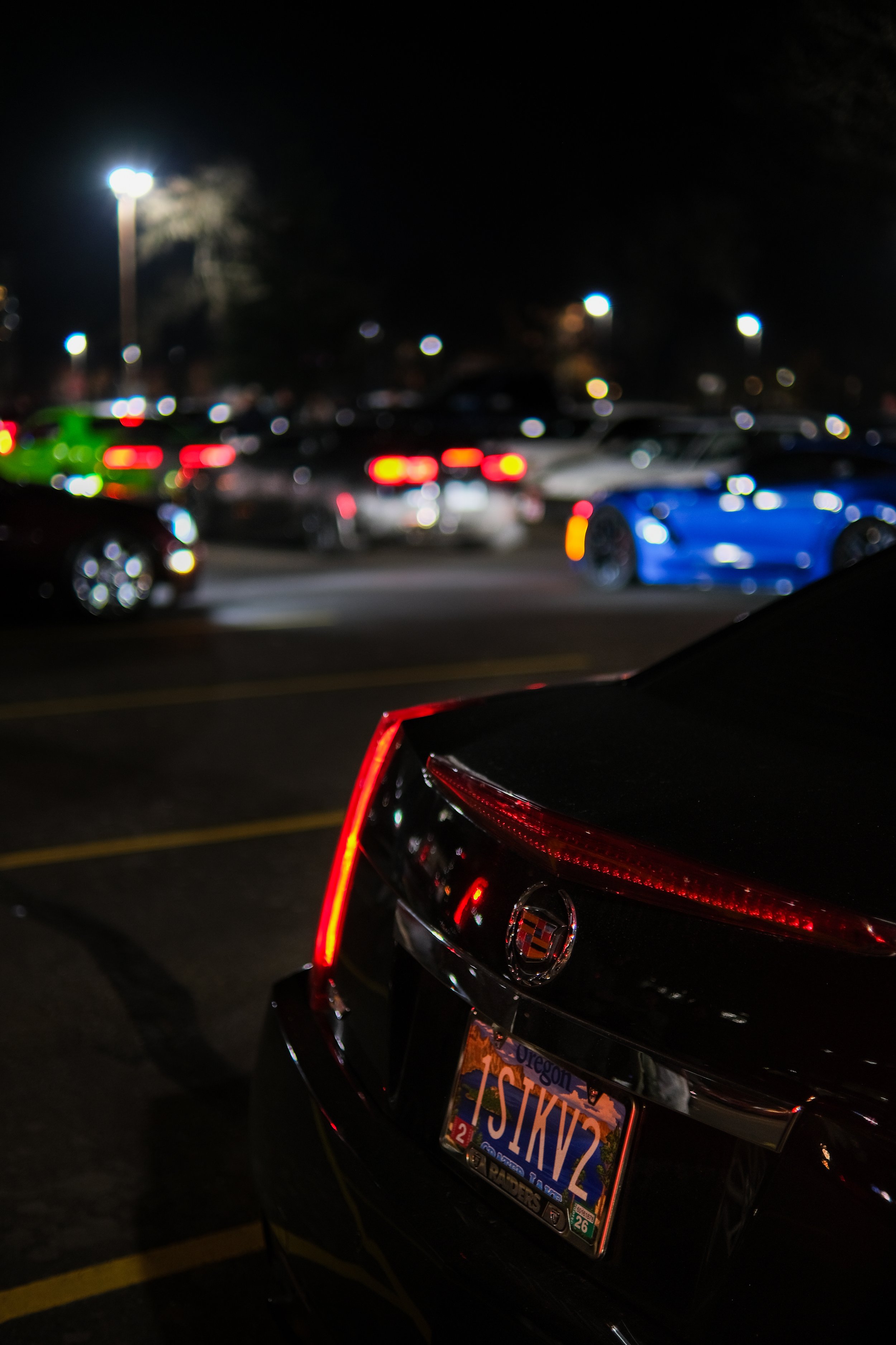 Close-up of a black Cadillac with an Oregon license plate parked in a busy parking lot at night, with other cars and bright lights in the background.