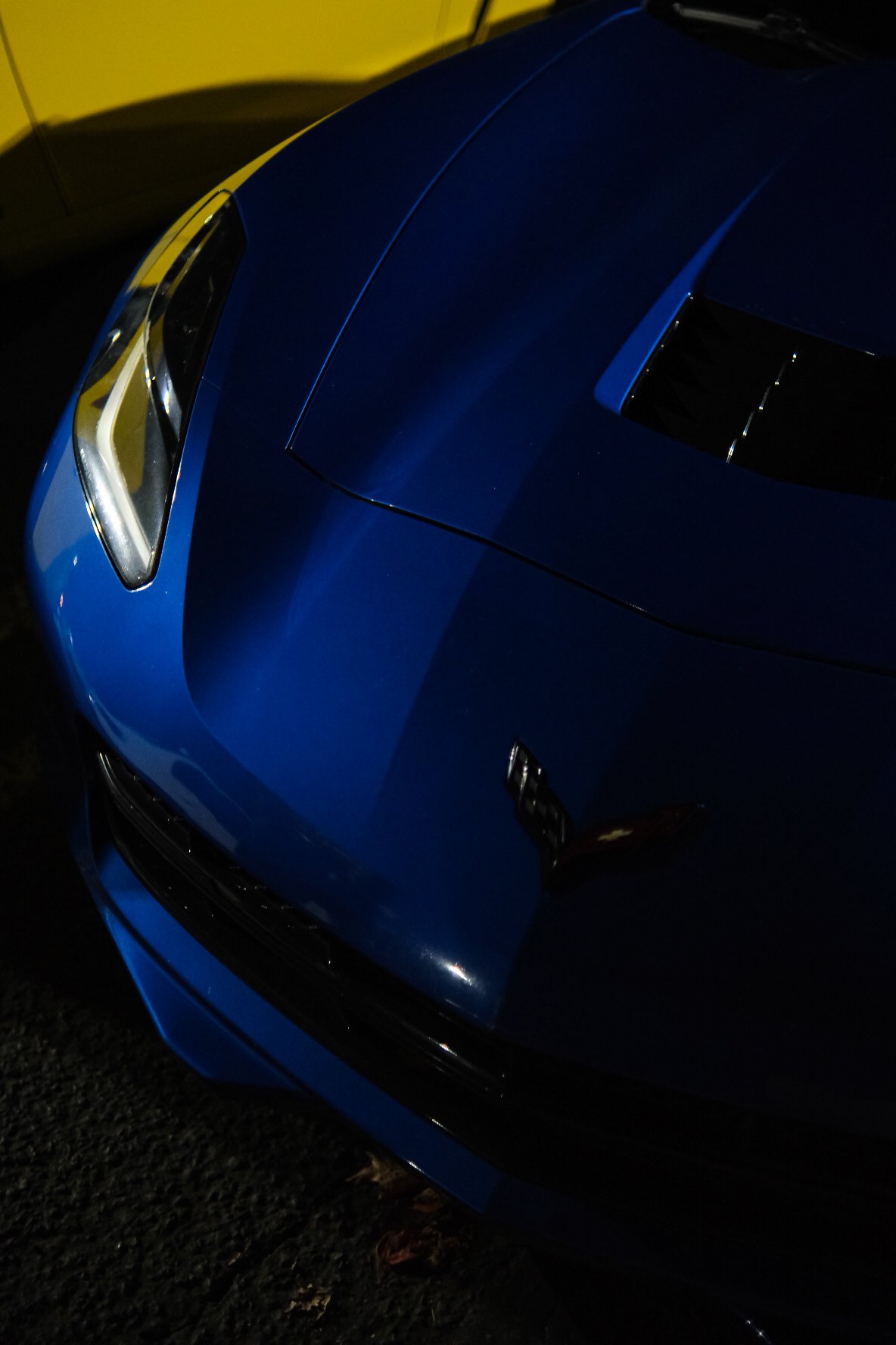 Close-up of the front end of a blue sports car, showing the headlight, hood, and part of the front grille, in a dimly lit environment.