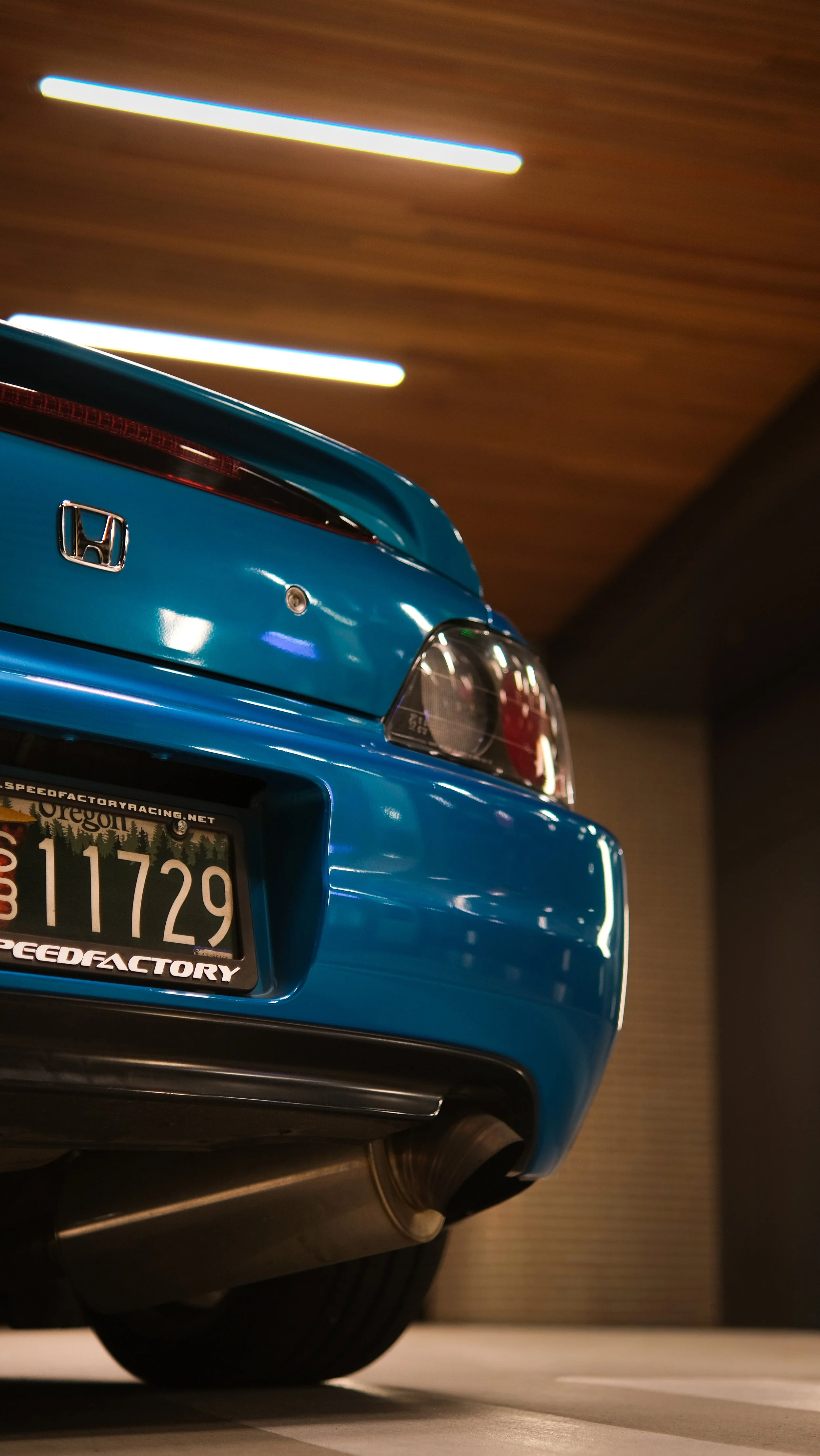 Close-up of the rear of a blue Honda car showing the back lights, rear bumper, exhaust pipe, and Oregon license plate with the number 11729, parked indoors with brown wood-paneled ceiling and ceiling lights.