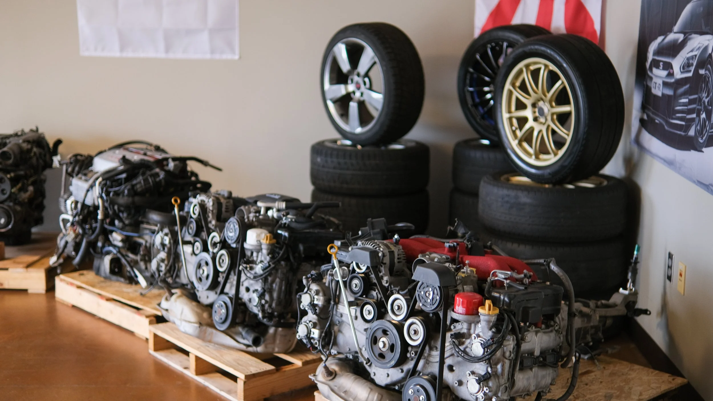 Several car engines on pallets and a display of car wheels mounted on stacks of tires to the right in a workshop or garage.