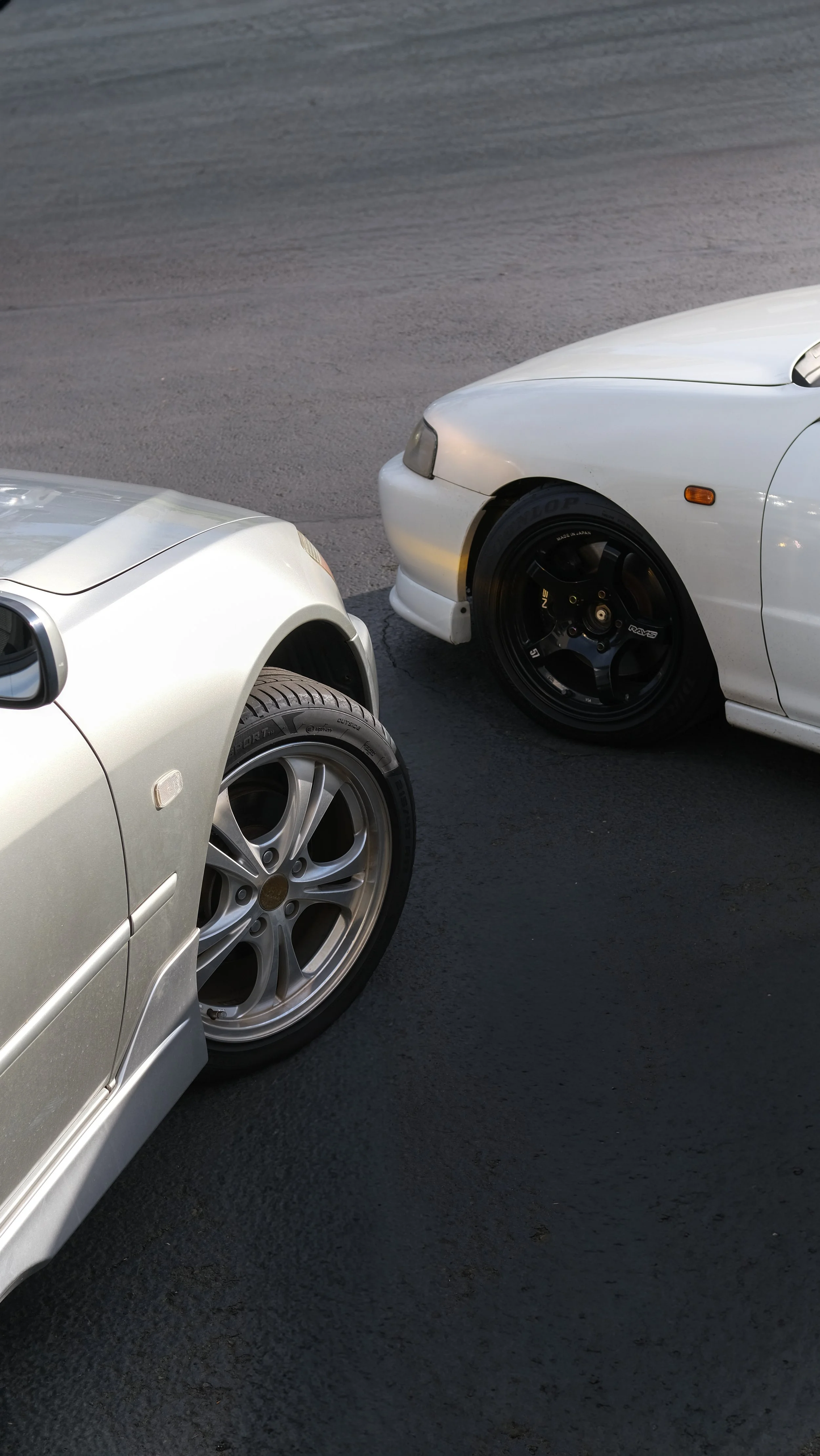 Close-up of two white sports cars parked on a paved surface, showing their front wheels and parts of the body, with one car slightly overlapping the other.