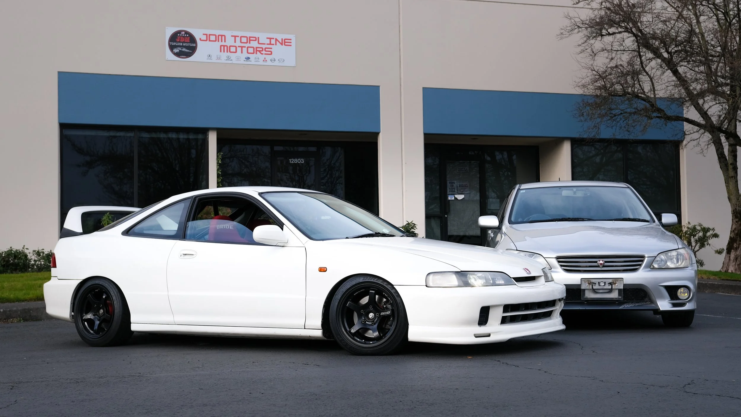 A white Honda Prelude with black wheels parked in front of a car dealership with a blue awning, alongside a silver car.