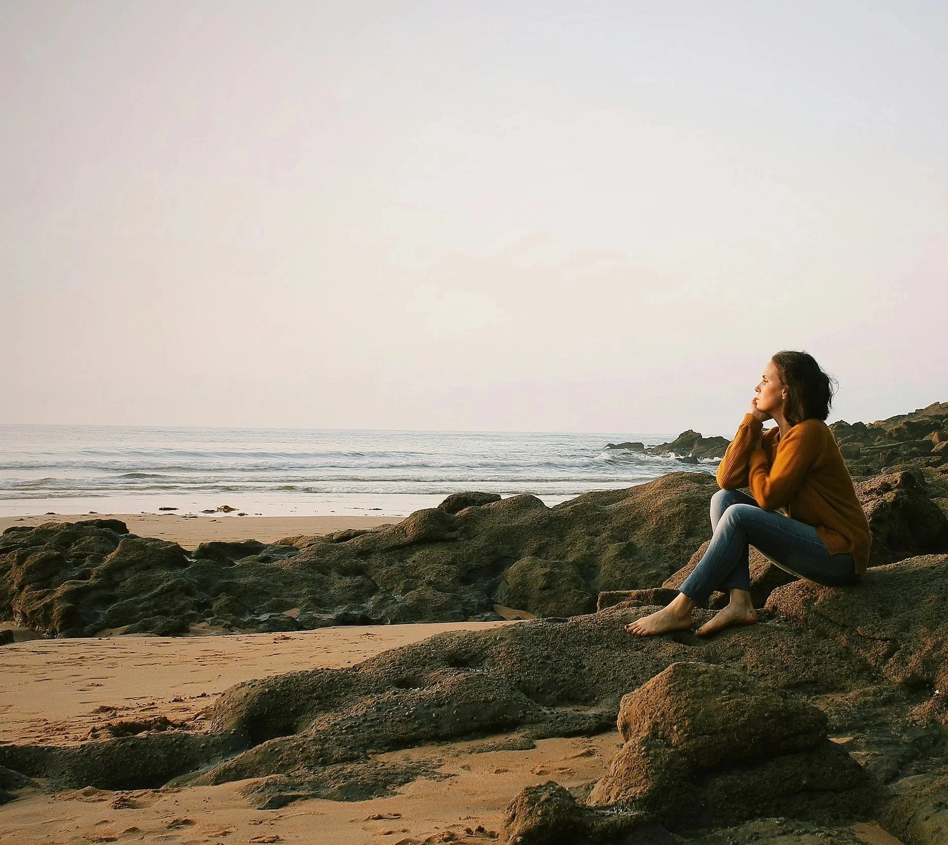 A woman sitting on rocks on a beach, looking out at the ocean during sunset or sunrise.