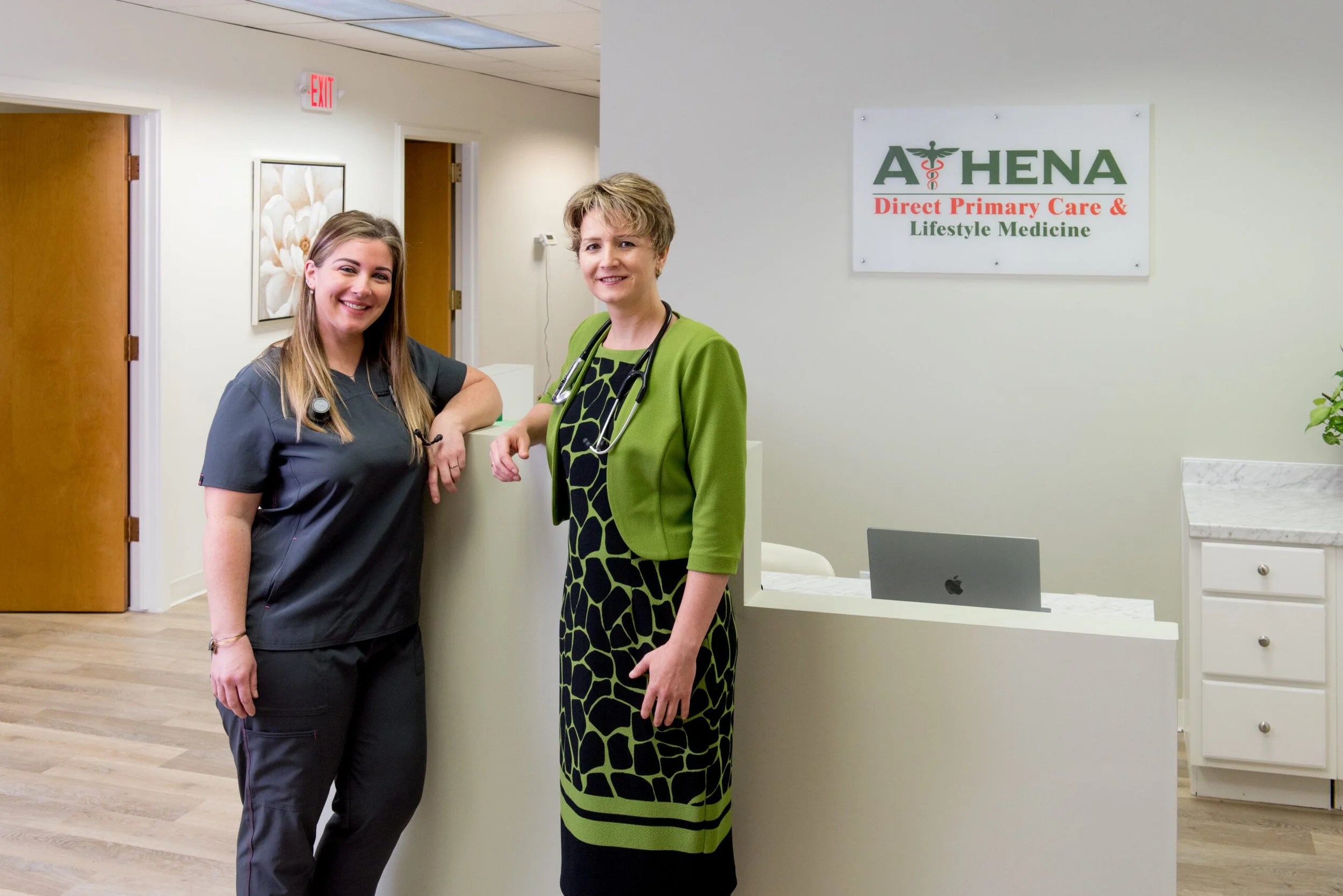 Two women, healthcare professionals, standing at the reception desk in a medical office, smiling. One is wearing black scrubs, the other a green cardigan with a patterned dress. Sign on the wall reads 'Athena Direct Primary Care & Lifestyle Medicine.'