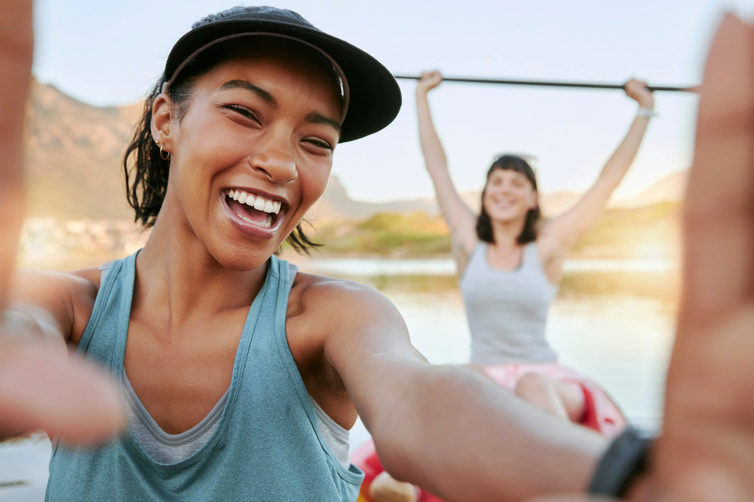 Two women, one in the foreground taking a selfie and smiling, and another in the background holding a paddle on a boat, with a scenic landscape and water behind them during sunset.