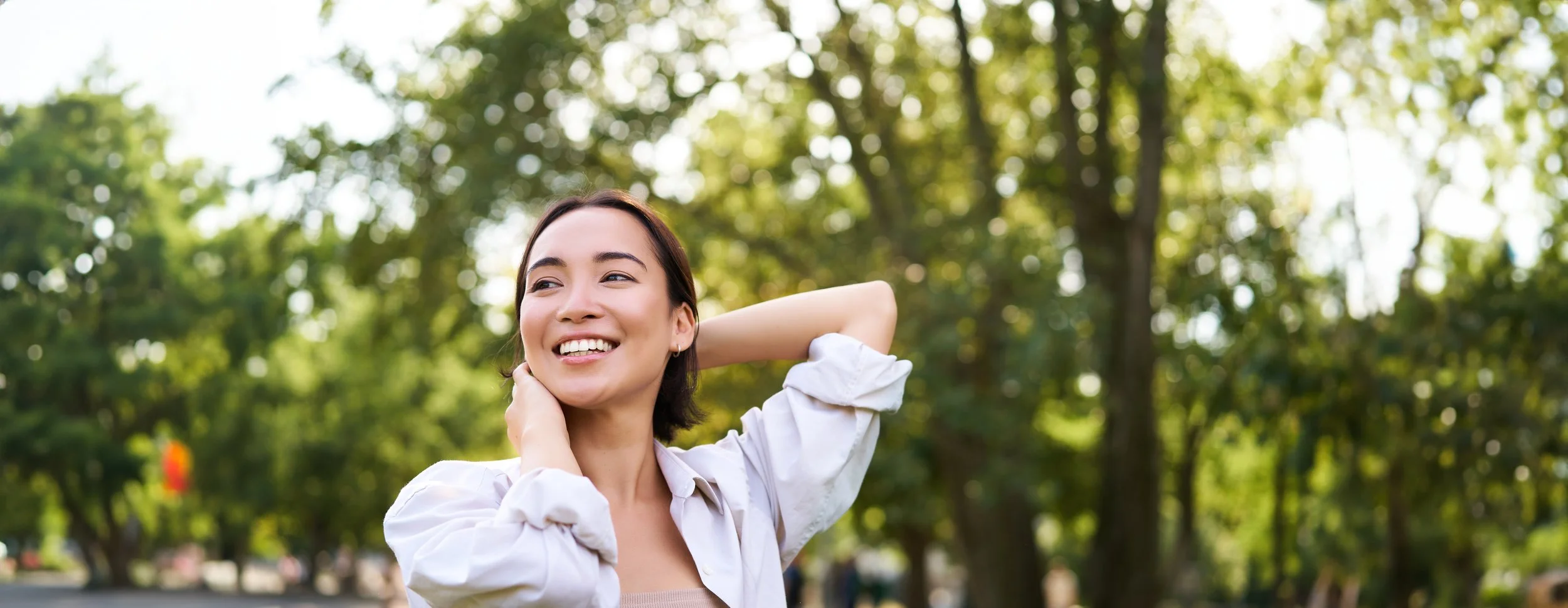 A woman smiling outdoors in a park with green trees in the background.
