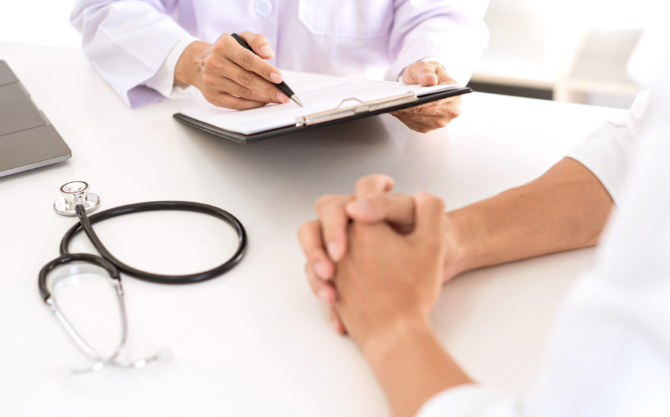 Doctor taking notes during consultation with patient, stethoscope on white table.