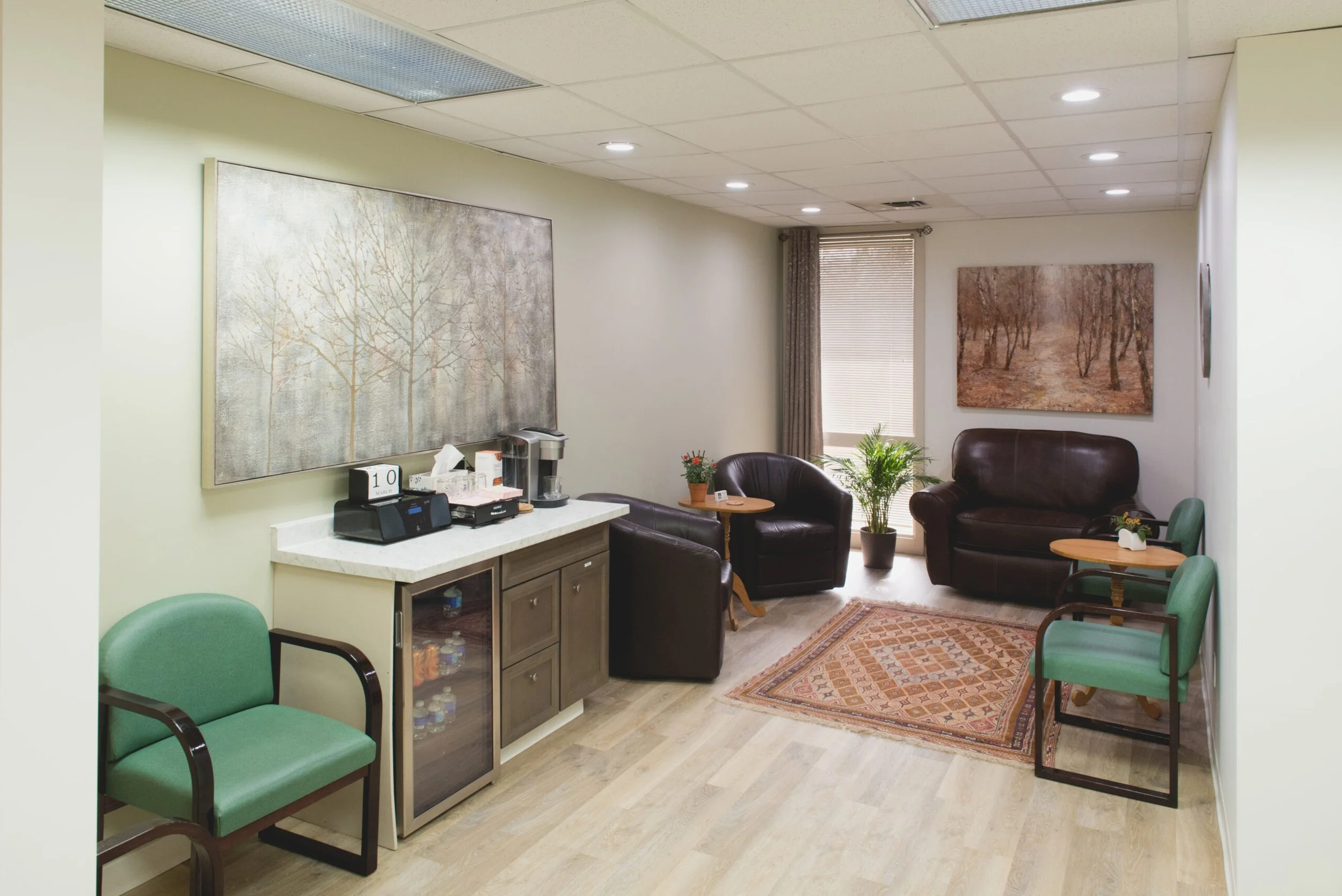 Comfortable waiting area with green and brown chairs, small tables, framed artwork of trees, a plant, and a rug on light wood flooring.