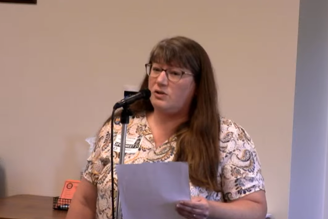 A woman with long brown hair, glasses, and a patterned shirt holding a paper and speaking into a microphone.