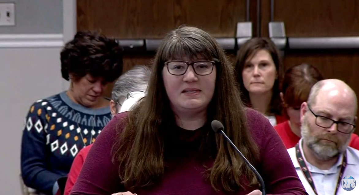 Woman with long brown hair and glasses speaking at a microphone during a panel or hearing, with several people seated behind her.