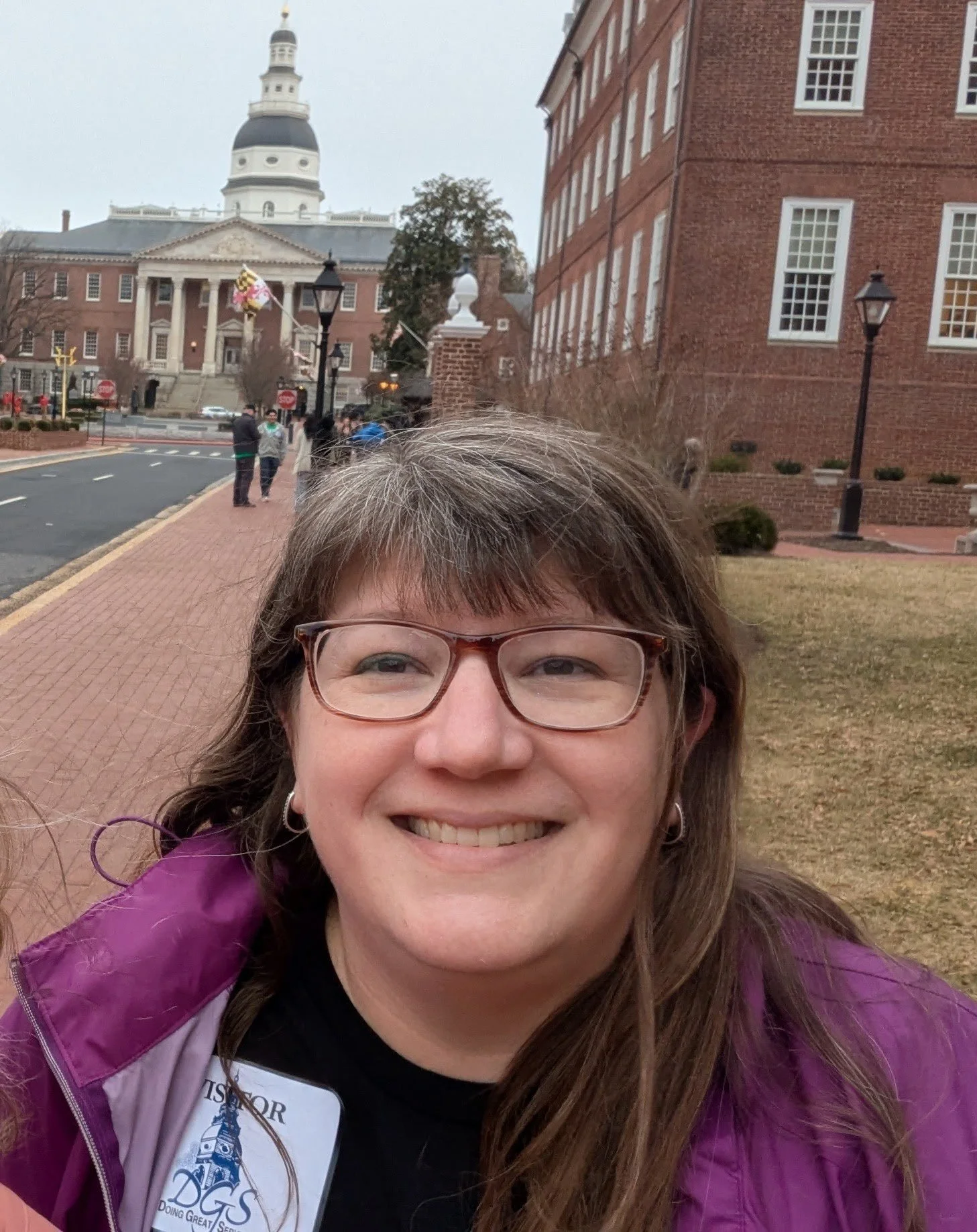 A woman with glasses and earrings smiling outdoors in front of a historic building with columns and a dome, with a brick sidewalk and other people in the background.