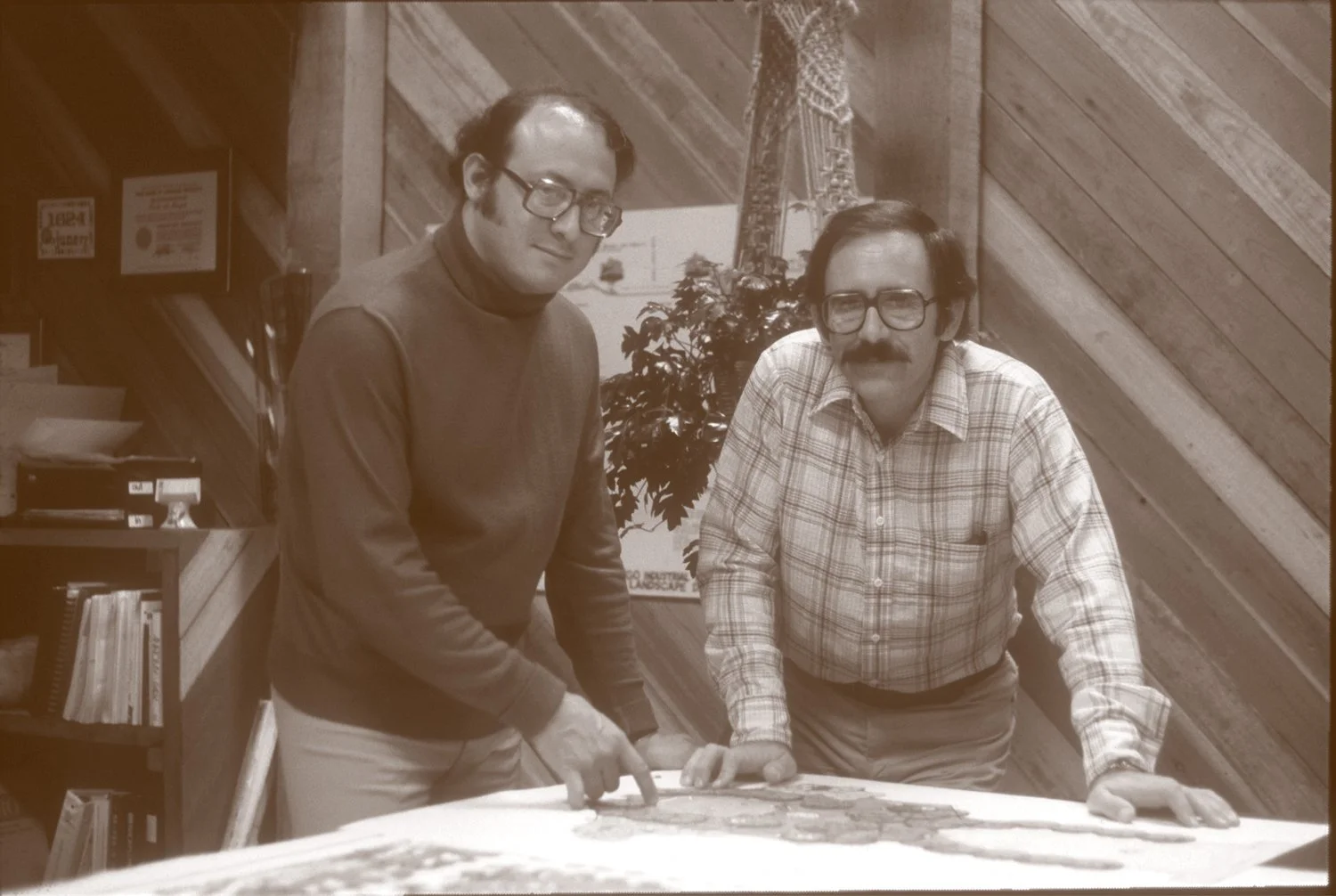 Retro image of Robert R. Cardoza, PLA, FASLA, and Peter DiLallo, PLA, ASLA, in 1971. Men with glasses and a mustache, wearing plaid and solid sweaters, are leaning over a table with a map or chart on it, in a room with wooden walls and bookshelves.