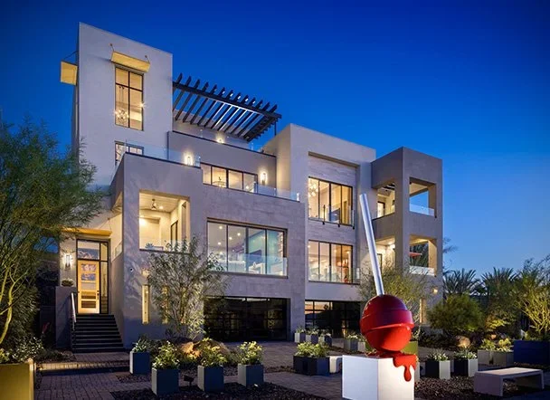 Modern multi-story house with large windows and outdoor lighting, decorated with a giant red Christmas ornament sculpture in the front yard.