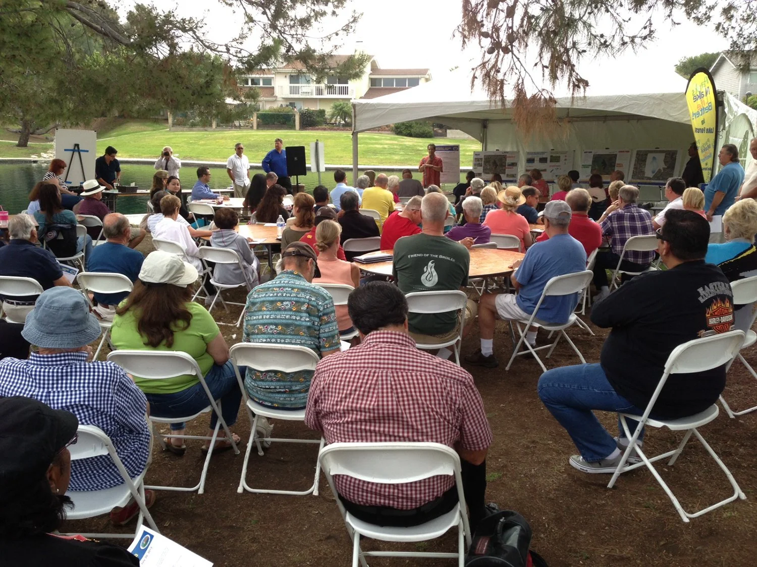 People attending an outdoor event or presentation under a white canopy near a pond, with trees and houses in the background.