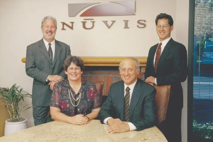 Four people, two men and two women, in business attire posing in an office with a sign that reads 'Nuvis' on the wall behind them. The woman seated at the table is wearing glasses and a patterned dress, while the man next to her is in a dark suit and tie. The man standing on the left has gray hair and a beard, and is wearing a gray suit. The man standing on the right has dark hair, glasses, and is also in a dark suit with a patterned tie.