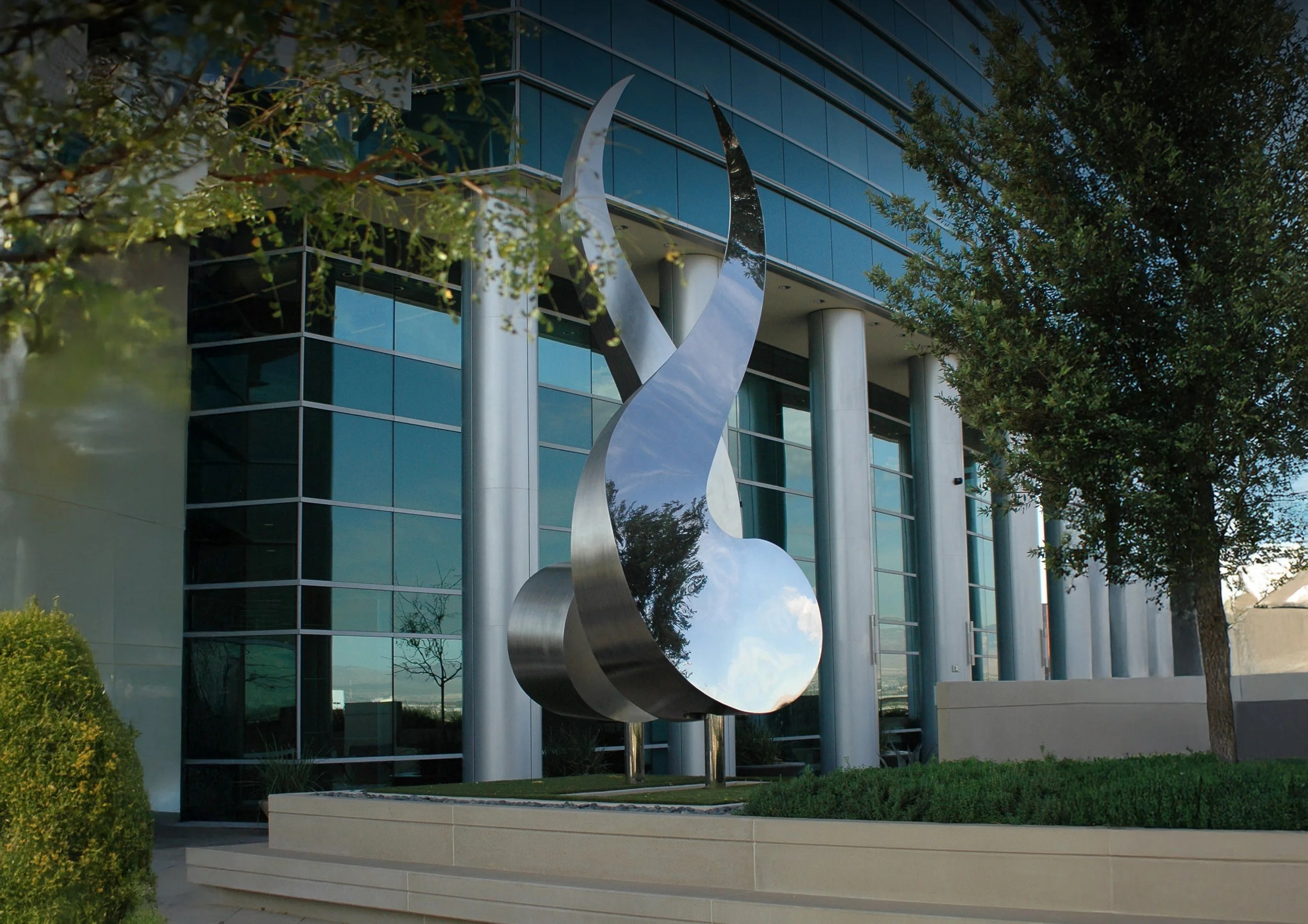 Modern office building with glass windows and a large metallic sculpture in front, surrounded by trees and greenery.