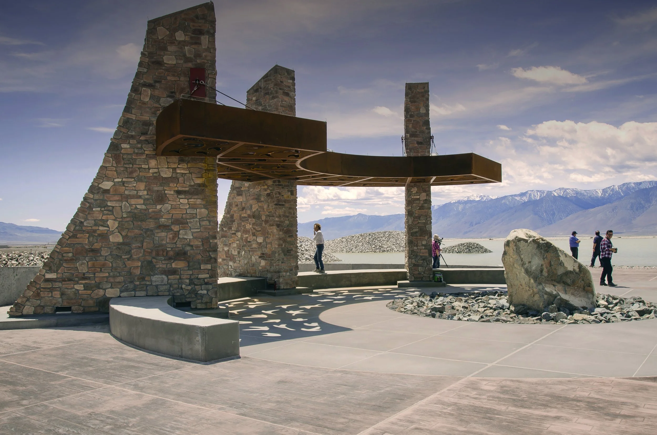 A brick and metal sculpture resembling a boat structure with three stone pillars, set against a backdrop of mountains, water, and a partly cloudy sky, with a few people standing nearby.