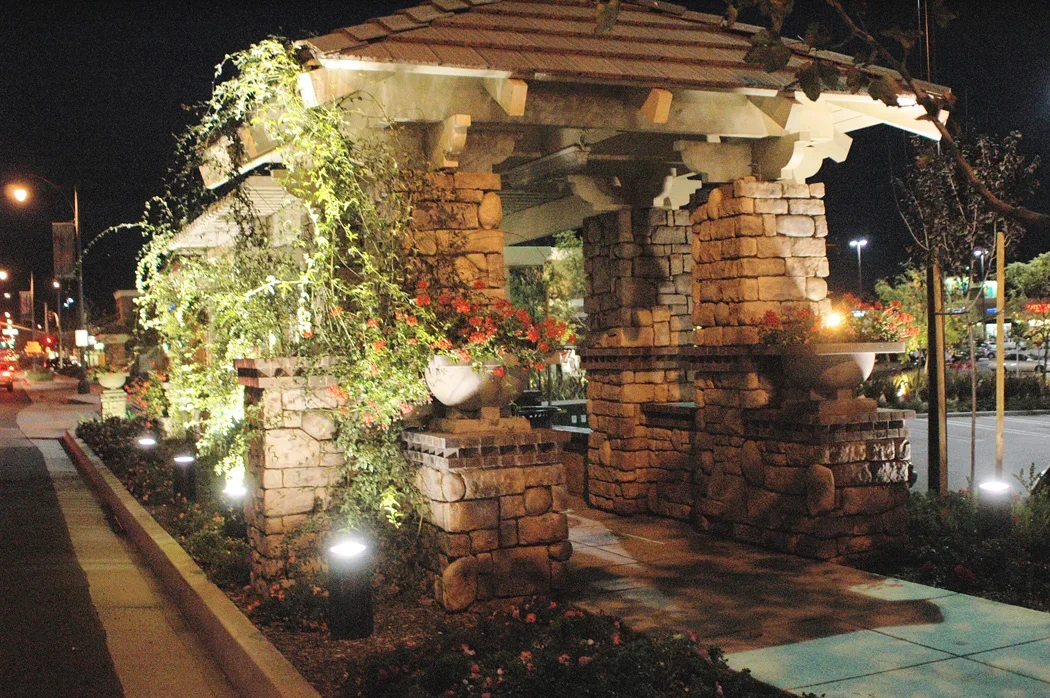 Night view of a stone pavilion with flower pots and plants, illuminated by landscape lighting, situated along a sidewalk in a commercial area.