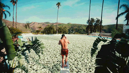 A man in swim trunks walking barefoot on a sandy beach toward the ocean. Tall palm trees are scattered along the beach with mountains in the background under a clear blue sky.